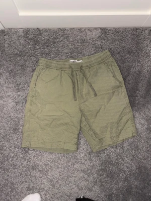 Olivgröna shorts från !Solid - Säljer ett par olivgröna shorts från !Solid med elastisk midja och snörning. Shortsen har två sidofickor och är tillverkade i ett lätt, strukturerat bomullstyg. Perfekta för varma dagar och enkel att matcha med t-shirt eller hoodie.