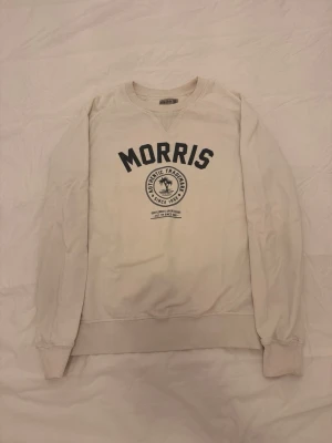 Vit sweatshirt från Morris - Klassisk vit sweatshirt från Morris med svart tryck framtill och rund halsringning. Tröjan har ribbade muddar vid ärmslut och nederkant samt en relaxed passform. Perfekt för en avslappnad och stilren look.