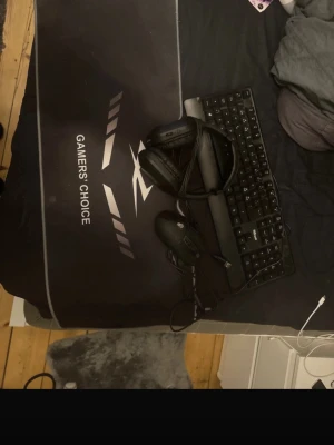 Gamingtillbehör paket Trust & musmatta - Säljer ett gamingpaket med svart Trust tangentbord, svart gamingmus, headset och en stor svart musmatta med texten 'Gamers' Choice'. Perfekt för dig som vill uppgradera din setup med snygga och funktionella tillbehör.