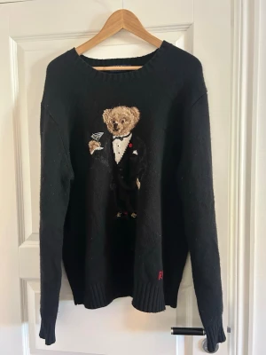 Svart stickad tröja Polo Ralph Lauren - Säljer Svart stickad tröja från Polo Ralph Lauren (äkta) med ikonisk björn i smoking och martiniglas broderad på framsidan. Tröjan har rund hals, ribbade muddar och är tillverkad i mjukt material. Perfekt för dig som gillar klassiska och lekfulla detaljer.