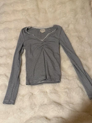 Randig långärmad topp Holly Whyte på Lindex  - Snygg långärmad topp från Holly Whyte på Lindex med smala svarta och vita ränder. Toppen har en v-ringning med vågig kant och rynkdetalj framtill. Tillverkad i mjukt och stretchigt material, perfekt för en avslappnad men trendig look. 