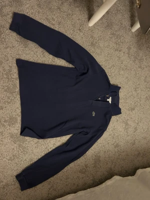 Mörkblå långärmad piké från Lacoste - Klassisk mörkblå långärmad pikétröja från Lacoste med den ikoniska krokodilloggan på bröstet. Tröjan har krage, knappar vid halsen och är tillverkad i mjukt bomullsmaterial. Perfekt för en clean och stilren look.