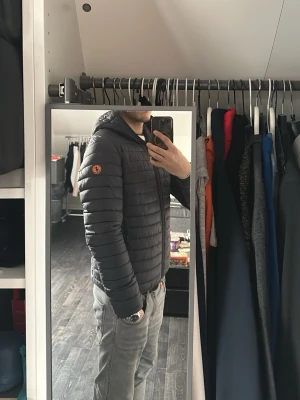 Svart lättviktsdunjacka från Save The Duck - Säljer en svart lättviktsdunjacka från Save The Duck med huva och dragkedja framtill. Jackan har en smal passform och diskret logotyp på ärmen. Perfekt för kalla vinterdagar och tillverkad i syntetiskt material utan animaliskt dun.