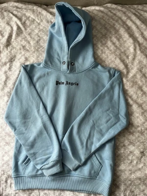 Ljusblå hoodie från Palm Angels - Snygg ljusblå hoodie från Palm Angels med svart logga på bröstet och stor text på ryggen. Hoodien har en stor huva med tryckknappar och klassisk magficka framtill. Tillverkad i mjukt bomullsmaterial och har ribbade muddar vid ärmslut och nederkant.