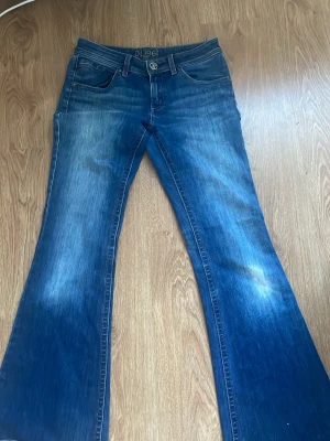 Blå utsvängda jeans med rå kant - Säljer ett par blå jeans från DL 1961 med utsvängda ben och rå, fransig kant längst ner. Jeansen har klassisk femficksmodell och snygga slitningar framtill och baktill. Perfekta för dig som vill sticka ut med en cool siluett.