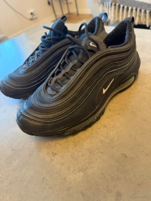 Nike Air Max 97 svarta sneakers - Säljer ett par nya oanvända svarta Nike Air Max 97 sneakers med ikoniska vågformade detaljer och synlig Air-sula. Skorna har en låg silhuett, snörning och små vita Swoosh-loggor på sidan och plösen. Materialet är en mix av textil och syntet.
