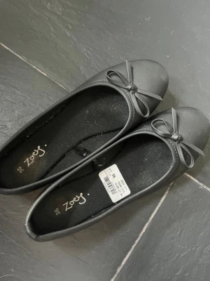 Svarta ballerinaskor med rosett  - Stilrena svarta ballerinaskor från Zoey med rund tå och dekorativ rosett framtill. Skorna har en klassisk, enkel design och är tillverkade i ett matt syntetmaterial. Perfekta för dig som gillar en clean och tidlös look. Storlek 36. Nypris 179kr. Endast använda två gånger.