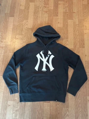 Svart New York Yankees hoodie - Snygg svart hoodie från New Era med stor vit New York Yankees-logga på bröstet. Klassisk känguruficka framtill och justerbar huva med dragsko. Tillverkad i mjuk bomullsblandning som är perfekt för chill dagar.
