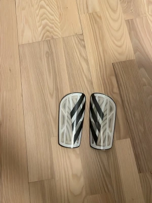 Vita benskydd från Adidas - Säljer ett par vita benskydd från Adidas med svarta ränder på framsidan. De har en lätt, böjd form och ventilerande mönster på insidan för extra komfort. Perfekta för fotbollsträning eller match.