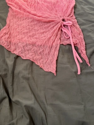 Rosa mesh topp med dragsko - Trendig rosa mesh topp med vågigt mönster och asymmetrisk nederkant. Toppen har en snygg dragsko med knytning framtill som ger en cool detalj. Materialet är tunt och lätt, perfekt för lager på lager eller som statement-plagg.