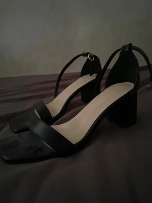 Svarta chunky sandaler med klack - Stilrena svarta sandaler med breda remmar och fyrkantig tå. Klackarna är chunky och ger både höjd och stabilitet. Justerbart spänne runt ankeln för perfekt passform. Ovandelen är i konstläder och innersulan är ljusbeige för en snygg kontrast.