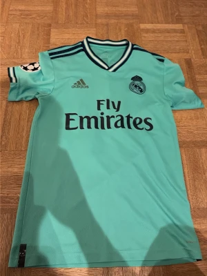 Real Madrid turkos matchtröja Adidas - Snygg turkos Real Madrid matchtröja från Adidas med svarta detaljer och tryck. Tröjan har klubbmärke, Adidas-logga och Fly Emirates på bröstet samt Champions League-märke på ärmen. På ryggen är det Lucas Vasquez och nummer 17. Perfekt för fotbollsfantaster och Real Supportrar som vill känna på känslan av lite halvt nostalgisk kärlek. Lite slitage gällande Champions märket och små flärparna nere vid tröjans slut, men även adidas märket är borta men texturen finns kvar. Annars funkar tröjan bra!