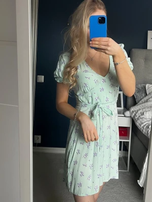 Mintgrön blommig klänning med knytband - Söt mintgrön klänning med små lila blommor, v-ringning och puffärmar. Midjeband som knyts för en snygg siluett. Lätt och luftig modell, perfekt för vår och sommar. Materialet ser ut att vara mjukt och bekvämt, troligen bomull eller viskos.