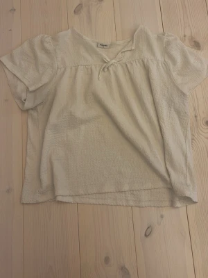 Beige blus med rosett från Kappahl - Söt beige blus från Kappahl i mjukt, strukturerat tyg. Blusen har korta puffärmar, v-ringning och en liten rosett framtill. Perfekt för dig som gillar en enkel men gullig stil.