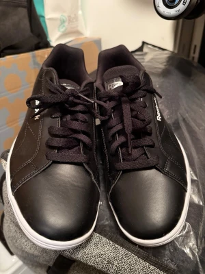 Reebok sneakers classic court clean 41 svarta - Snygga svarta sneakers från Reebok med klassisk låg profil. Skorna har ovandel i slätt läder, svarta skosnören och vit sula. Reebok-logga i vitt på sidan. Perfekta för en clean och stilren look.