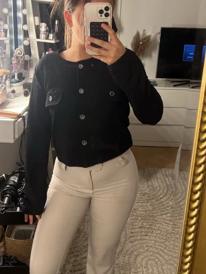 Svart cardigan med glittriga knappar - Trendig svart cardigan med rund hals och glittriga, dekorativa knappar framtill. Kortare modell med två små fickor på bröstet. Perfekt att styla till både jeans och kjol