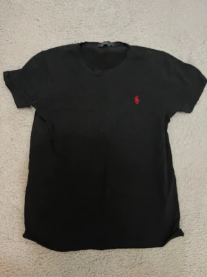 Svart t-shirt från Polo Ralph Lauren - Klassisk svart t-shirt från Polo Ralph Lauren med röd broderad logga på bröstet. T-shirten har rund halsringning och korta ärmar. Tillverkad i mjuk bomull för en skön känsla och enkel stil.