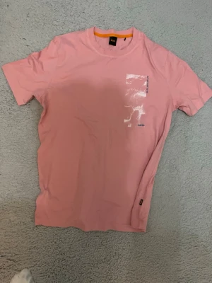 Rosa t-shirt från BOSS - Snygg rosa t-shirt från BOSS med korta ärmar och rund hals. Framtill finns en blank, rektangulär detalj och en liten BOSS-logga. Perfekt för dig som gillar stilrena plagg med en twist.
