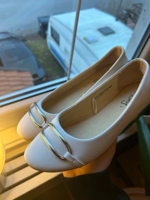 Vita ballerinaskor med metallspänne - Stilrena vita ballerinaskor med rund tå och dekorativt metallspänne framtill. Skorna har en enkel och klassisk design som passar till många olika outfits. Tillverkade i ett slätt material med mjuk innersula för extra komfort.