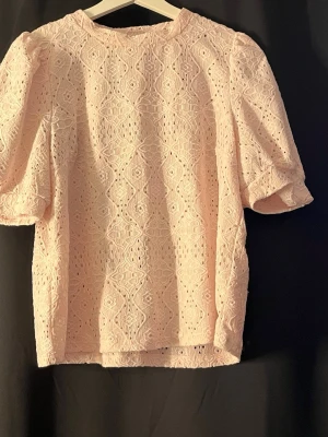 Ljusrosa spetsblus med puffärm - Supersöt ljusrosa blus med puffärmar och vackert spetsmönster. Blusen har rund halsringning och är perfekt för dig som gillar romantiska detaljer. Materialet är mjukt och luftigt, vilket gör den skön att bära under varmare dagar.