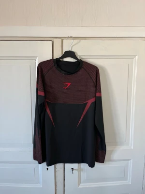 Gymshark Onyx V5 Long sleeve - Snygg långärmad träningströja från Gymshark i svart med röda detaljer och mönstrade partier på axlar och ärmar. Tröjan har en sportig design med rund hals och är tillverkad i ett stretchigt funktionsmaterial som passar perfekt till gymmet eller löpning.