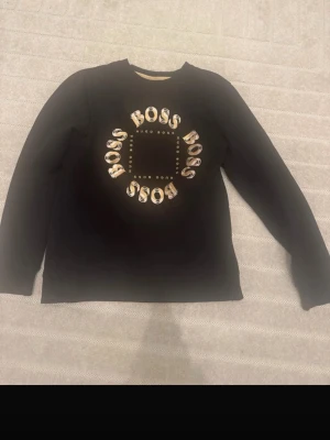 Svart sweatshirt från Hugo Boss - Snygg svart sweatshirt från Hugo Boss med stort cirkelformat BOSS-tryck i guld och vitt på bröstet. Tröjan har rund halsringning och långa ärmar. Perfekt för dig som gillar streetwear och vill ha en tröja med tydlig logga.