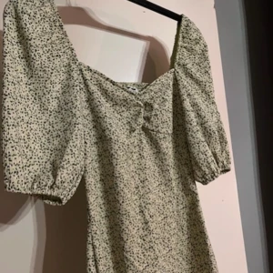 Ljus blommig kortklänning med puffärm - Supersöt ljus kortklänning med småblommigt mönster och puffärmar. Klänningen har v-ringning och en liten rosett framtill. Perfekt för sommarens alla tillfällen och ger en romantisk vibe.