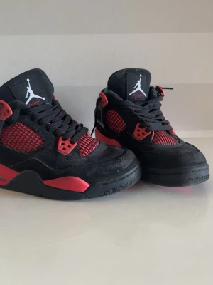 Svarta och röda Air Jordan 4 sneakers - Säljer ett par Air Jordan 4 sneakers i svart och rött med klassisk Jumpman-logga på plösen. Skorna har meshdetaljer på sidorna, svarta skosnören och röda accenter på sulan och snörningen. Ovandelen är i mocka och textil för en sportig look.