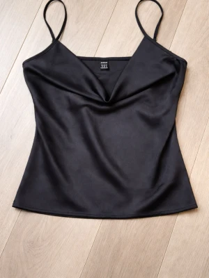 Svart satinlinnetopp med smala band - waterfall - Snygg svart linnetopp i glansigt satinmaterial. Toppen har smala axelband och en lätt draperad v-ringning som ger en elegant look. Perfekt att styla med jeans eller kjol för en trendig outfit. Liten i storlek 