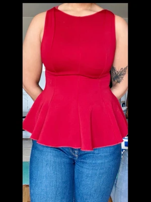 Röd peplum topp utan ärmar - Snygg röd peplum topp med figurnära passform och utsvängd nederdel. Toppen är ärmlös och har en hög halsringning som ger en clean look. Perfekt att styla med jeans eller kjol för en trendig outfit. Använt en gång 