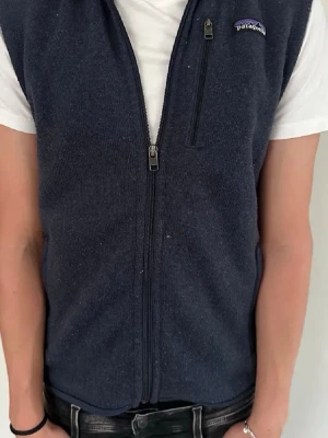 Mörkblå fleeceväst från Patagonia - Säljer en mörkblå fleeceväst från Patagonia med hel dragkedja framtill och en bröstficka med dragkedja. Västen har en enkel och stilren design, perfekt att slänga över en t-shirt för extra värme. Klassisk logga på bröstet.