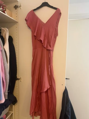 MörkRosa omlottklänning med volang strl 38 - Säljer en lång, rosa omlottklänning med v-ringning och volangdetaljer framtill. Klänningen har ett mjukt, flytande tyg och är ärmlös med en elegant, asymmetrisk skärning. Perfekt för sommarens alla tillfällen! Strl 38. Mörkrosa. Använd en gång. Köpt på Boozt.  Märke Minus 