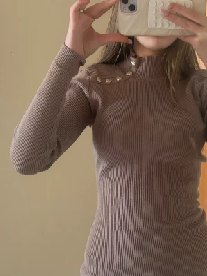 Beige ribbad polotröja med knappar - Trendig beige ribbad polotröja med långa ärmar och dekorativa pärlemorknappar på axeln. Figurnära passform och hög hals ger en clean och stilren look. Perfekt att matcha med jeans eller kjol för en snygg outfit.