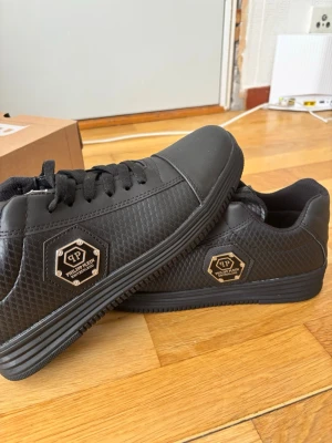 Svarta sneakers från Philipp Plein - Snygga svarta sneakers från Philipp Plein med hexagonformad metall-logga på sidan. Skorna har en låg profil, klassiska svarta skosnören och en texturerad yta i syntetmaterial. Perfekta för dig som vill ha en stilren och modern look.
