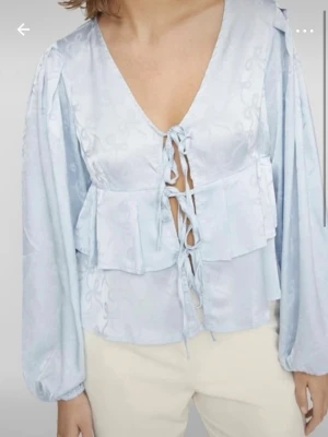Ljusblå blus med knytning fram - Trendig ljusblå blus i satinliknande material med v-ringning och dekorativa knytningar framtill. Blusen har volangdetaljer vid midjan och långa, puffiga ärmar med resår vid ärmslut. Perfekt för en stilren och modern look.