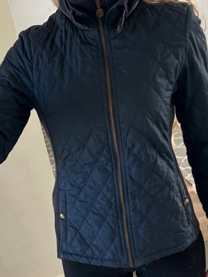 ommy Hilfiger quiltad jacka  - Snygg marinblå quiltad jacka med dragkedja framtill och hög krage. Jackan har ribbade partier på sidorna, två fickor med tryckknappar och ett emblem på ärmen. Perfekt för dig som vill ha en stilren och bekväm jacka till vardags. 