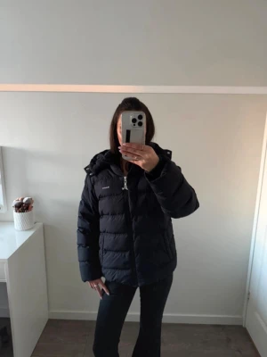 GANT pufferjacka - Säljer en marinblå pufferjacka från GANT med huva och dragkedja framtill. Jackan har en klassisk quiltad design och logotyp på bröstet. 