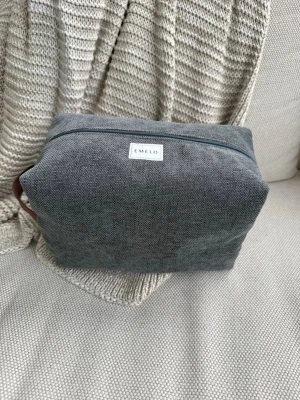 Grå necessär från Emelo i canvas - Snygg och stilren grå necessär från Emelo i tåligt möbeltyg. Väskan har en rektangulär form, dragkedja upptill och ett brunt handtag i sidan. Perfekt för att organisera småprylar eller smink i väskan. Brunt innertyg. Mått ca. 26x17x12 cm