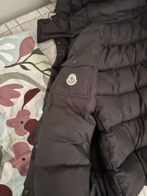 Svart dunjacka från Moncler - Säljer en svart dunjacka från Moncler med klassisk quiltad design och stor huva. Jackan har flera fickor med knappar och Moncler-logga på ärmen. Perfekt för kalla vinterdagar och riktigt snygg att bära till jeans eller joggers.