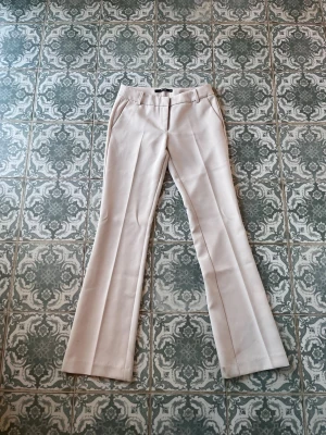 Beige kostymbyxor med pressveck - Snygga beige kostymbyxor med pressveck och utsvängda ben. Byxorna har fickor fram, hällor för skärp och dekorfickor bak. Perfekta för en clean och stilren look. Materialet känns mjukt och har en lätt stretch för extra komfort.