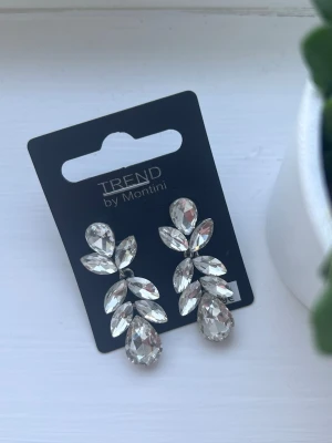 Silvriga statementörhängen Montini - Glittriga örhängen från Montini med stora, klara stenar i dropp- och bladform. Örhängena har en silvrig metallbas och ger en riktigt lyxig känsla. 