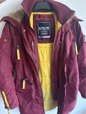 Vinröd Alpha Industries parkas - Snygg vinröd parkas från Alpha Industries. Är i använt skick och har lite missfärgningar på ärmarna och lite på jackan. Jackan är i strl M men passar mig som vanligtvis bär XS-S