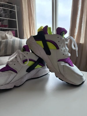 Nike Air Huarache vita/lila/gula - Nike Air Huarache sneakers i vitt med detaljer i lila, neon-gult och svart. Skorna har en chunky sula, klassisk Huarache-plastdetalj vid hälen och ventilerande meshpartier. Perfekt för dig som gillar retrostil och färgstarka sneakers. Storlek 40,5 men passar bättre storlek 39.