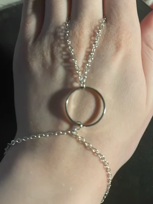 Silvrigt handkedjearmband med ring - Snyggt handkedjearmband i silverfärgad metall med en enkel cirkelformad ring på ovansidan av handen. Kedjan går från handleden till fingret och ger en modern och stilren look. Perfekt accessoar för att lyfta din outfit.OBS! Materialet är okänt, kan ej garantera att det är rostfritt.
