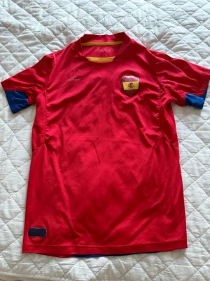 Spaniens röd supportertröja - Röd officiell supportertröja för Spaniens landslag från soc. Tröjan har blå detaljer på ärmarna, gult inslag vid kragen och ett broderat landslagsemblem på bröstet. Tillverkad i lätt och ventilerande material, perfekt för fotboll och sport. Bedöm skicket själva