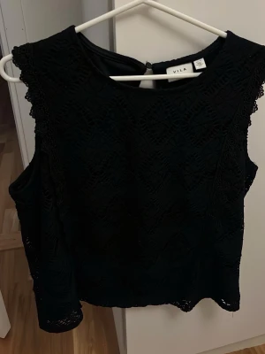 Svart spetsblus från Vila - Säljer en svart blus från Vila med vackra spetsdetaljer och mönstrad struktur. Blusen är ärmlös och har en rund halsringning. Perfekt för dig som gillar en stilren men ändå detaljerad look.