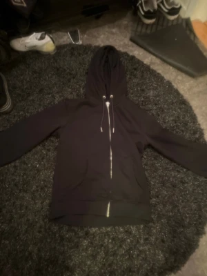 Svart zip hoodie med dragsko - En stilren svart hoodie med dragkedja framtill och justerbar dragsko i huvan. Hoodien har två stora fickor på magen och ribbade muddar vid ärmslut och nederkant. Perfekt för en avslappnad och cool look.