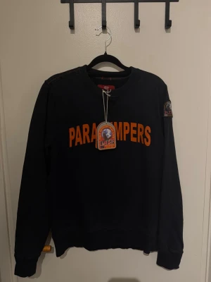 Svart sweatshirt från Parajumpers - Cool svart sweatshirt från Parajumpers med orange text på bröstet och patch på ärmen. Klassisk rund halsringning och ribbade muddar. Perfekt för en avslappnad streetwear-look.