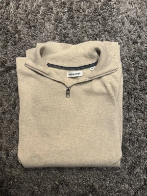 Beige stickad tröja Jack & Jones - Snygg beige stickad tröja från Jack & Jones med halv dragkedja vid halsen och ribbade muddar. Tröjan har en stilren design och är perfekt för lager på lager. Materialet känns mjukt och bekvämt, och färgen är lätt att matcha med olika outfits.