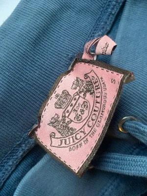 Blå velourbyxor Juicy Couture - Mysiga blå mjukisbyxor från Juicy Couture i klassisk velour. Byxorna har dragsko i midjan och breda muddar. Perfekta för en chill och avslappnad stil. Ikonisk rosa etikett med logga på insidan.